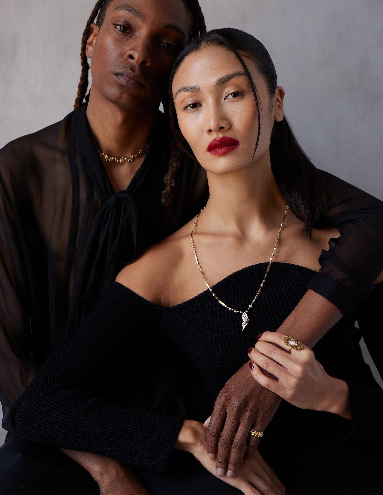 A man and a woman sitting close, wearing black clothing. The woman is showcasing a pinky ring with a textured effect.