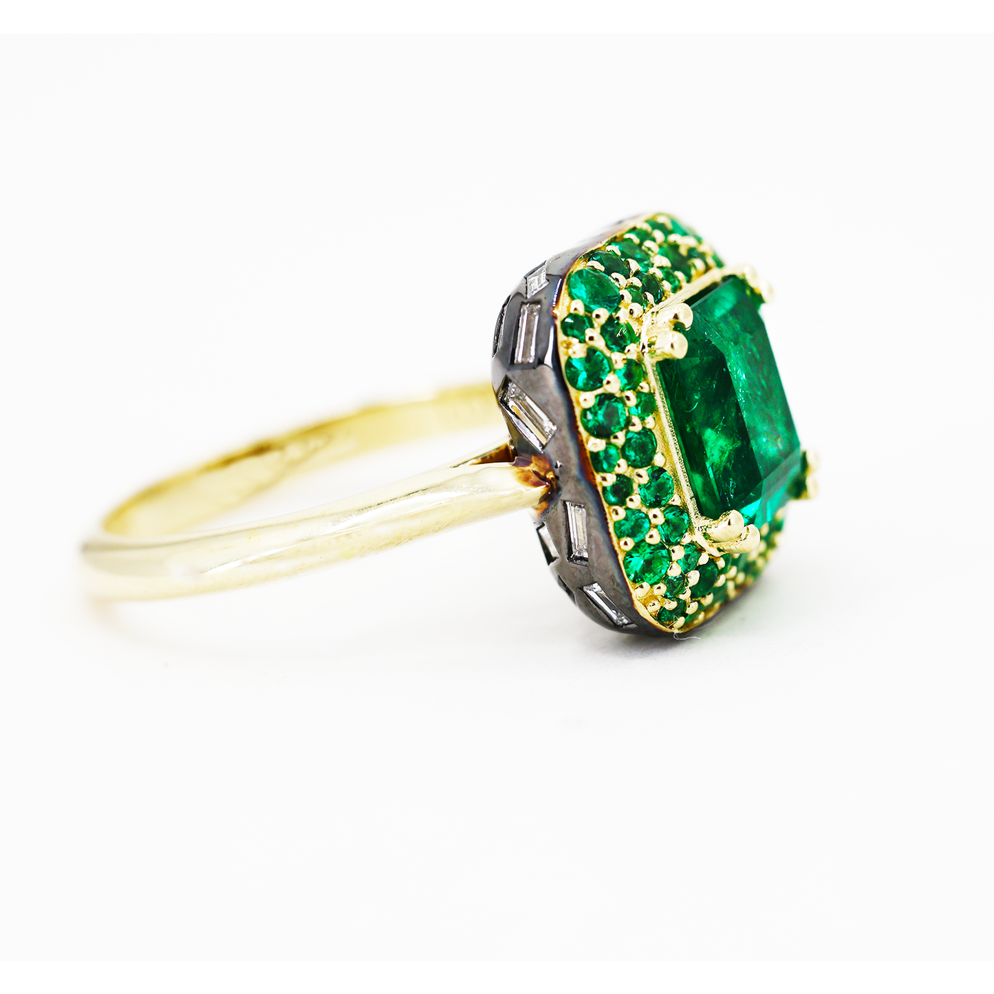An elegant gold ring featuring an octagon cut Zambian natural 2ct emerald on a domed halo with pave emeralds and baguette natural diamonds set on the underside, with a 2mm band swooping up to ridged shoulders.