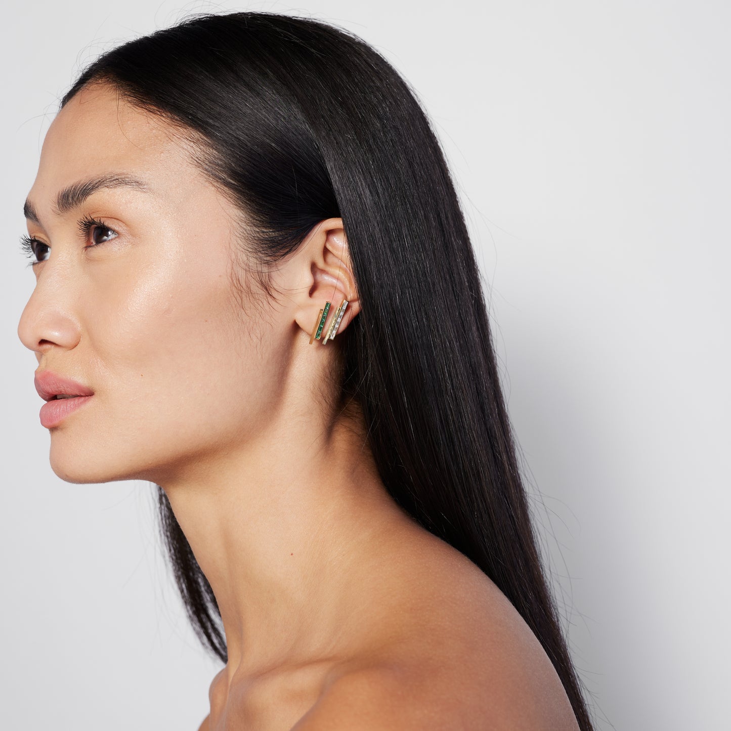 A model wearing a pair of gold stud earrings with pinstripe bar design set with either diamonds or emeralds.