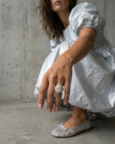 Person wearing a white dress and large diamond ring with puffed sleeves, sitting on a concrete floor.