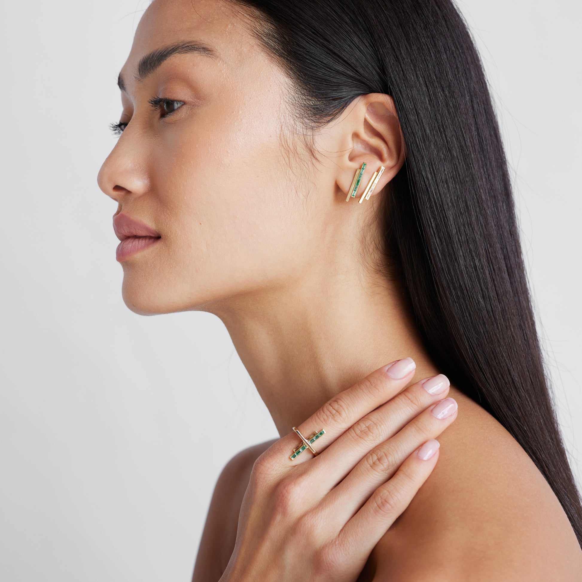 A model wearing a pair of gold stud earrings with pinstripe bar design set with either diamonds or emeralds.