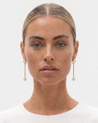 A close-up portrait of a woman wearing long, delicate gold dangle earrings with small triangular charms with diamonds