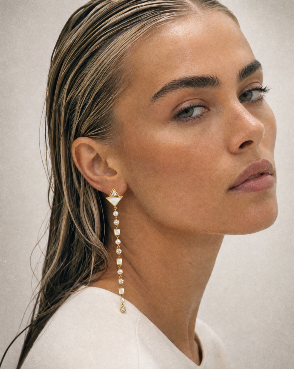 A close-up portrait of a woman with wet, styled hair wearing a long, dangling gold and pearl earring, set against a neutral light gray background.
