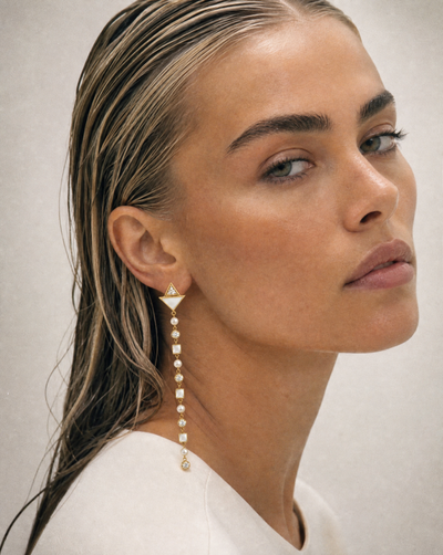 A close-up portrait of a woman with wet, styled hair wearing a long, dangling gold and pearl earring, set against a neutral light gray background.
